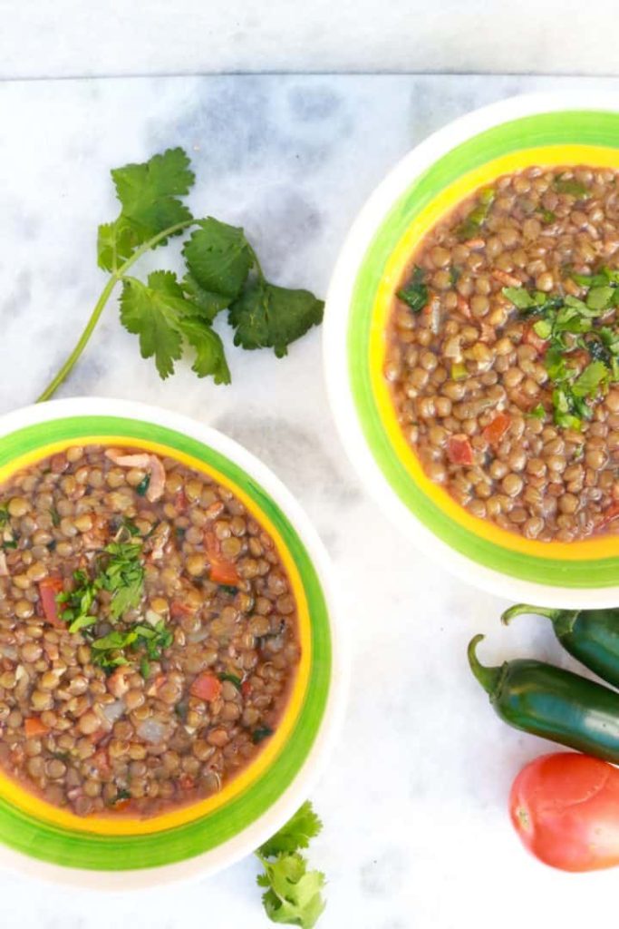Two bowls of Mexican Lentil Soup with cilantro, jalapeños and tomatoes around them