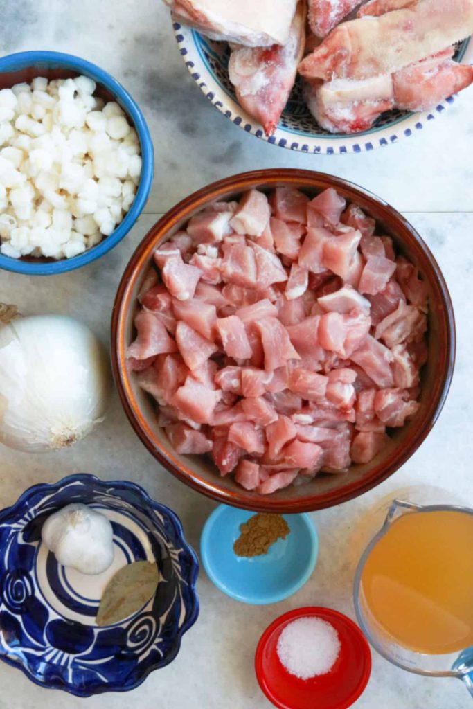 Bowl with raw, diced pork, bowl of pig's feet, hominy, onion, garlic, bay leaf, cumin, salt and broth