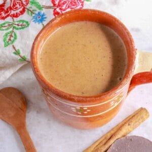 A warm mug of Mexican champurrado with a cinnamon stick and Mexican chocolate disc beside it.