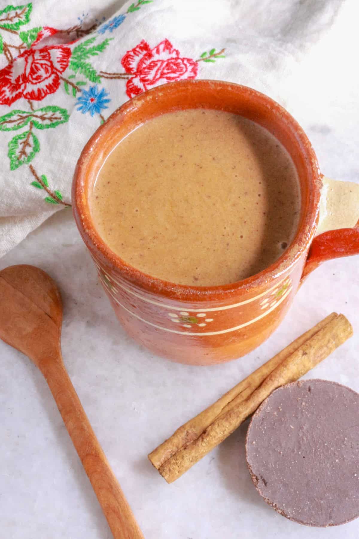 A warm mug of Mexican champurrado with a cinnamon stick and Mexican chocolate disc beside it.