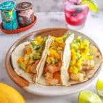 A plate with three shrimp tacos with mango salsa and a sparkling blueberry mocktail behind it