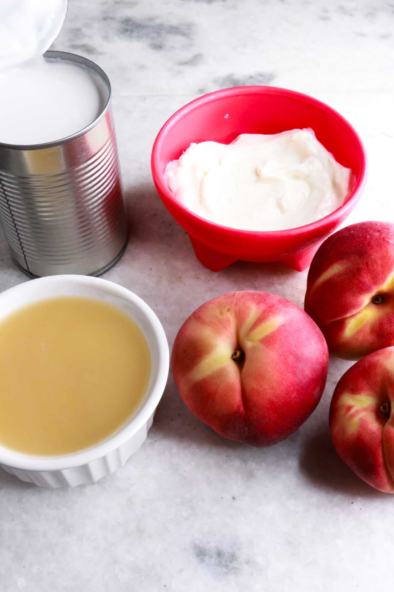Can of coconut cream, a bowl with yogurt, a bowl with coconut condensed milk, and 3 fresh peaches