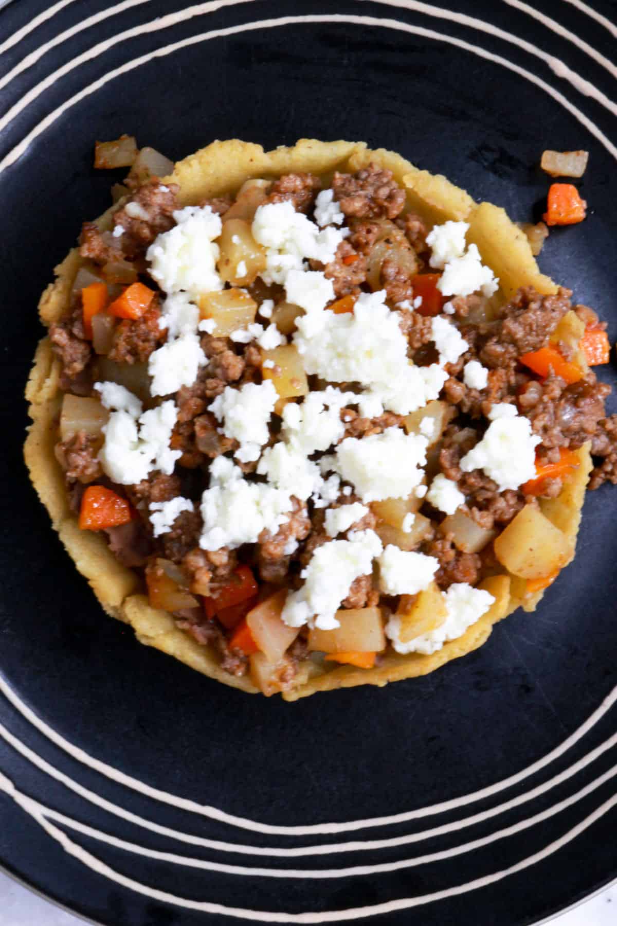 A sope with beans, picadillo and queso fresco on a black plate