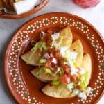 Three tacos dorados de pollo on a brown Mexican plate topped with garnishes