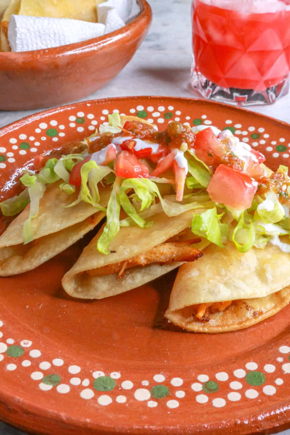 Three tacos dorados de pollo (crispy chicken tacos) on a Mexican plate, topped with garnishes