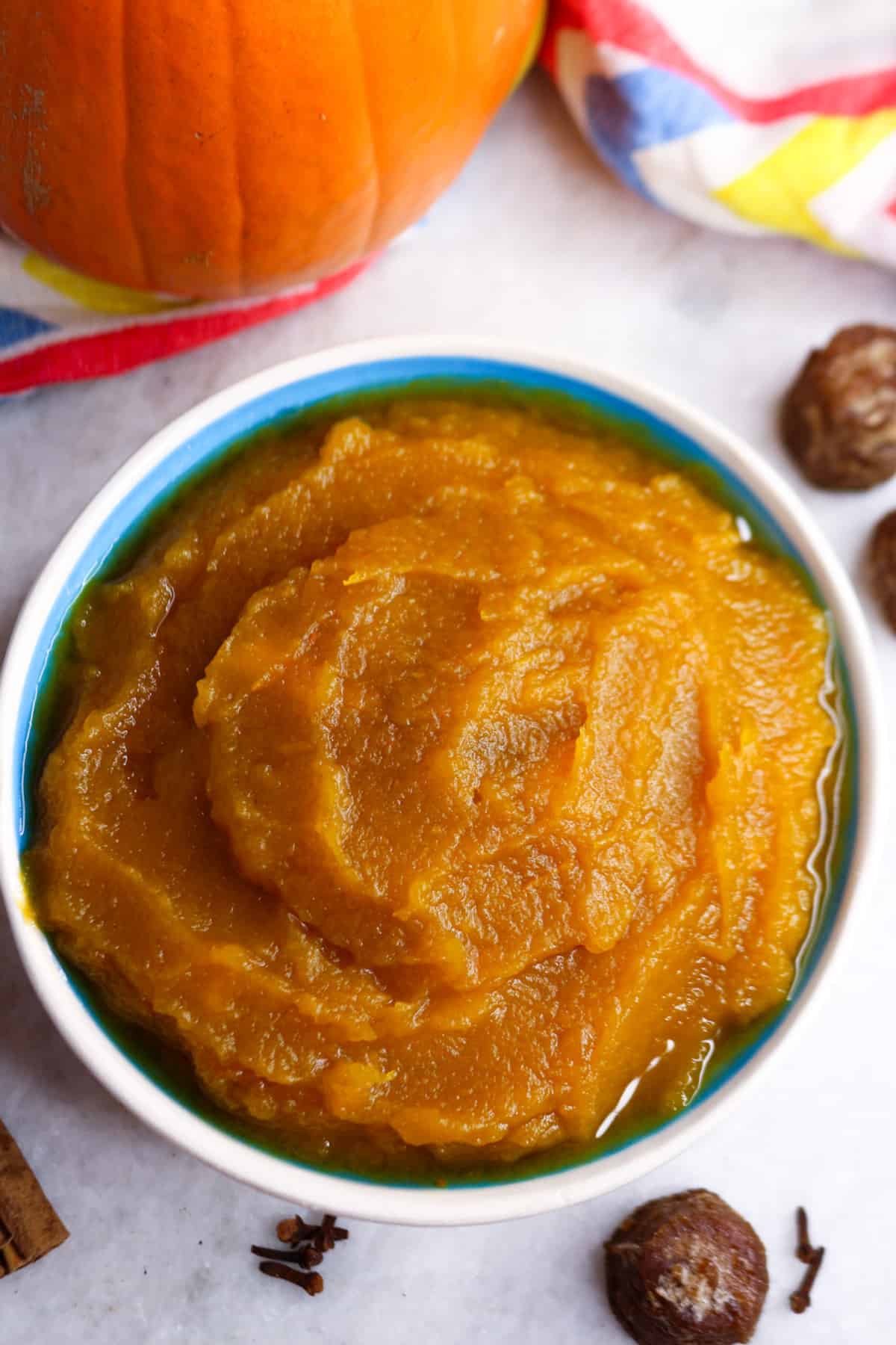 A bowl of puré de calabaza in a bowl surrounded by small piloncillos, a pumpkin, cinnamon stick and cloves