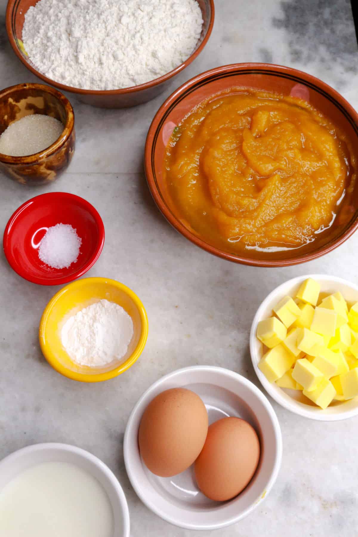 A bold of pumpkin puree, a bowl of gluten-free flour, sugar, baking powder, salt, two eggs, butter in cubes and milk