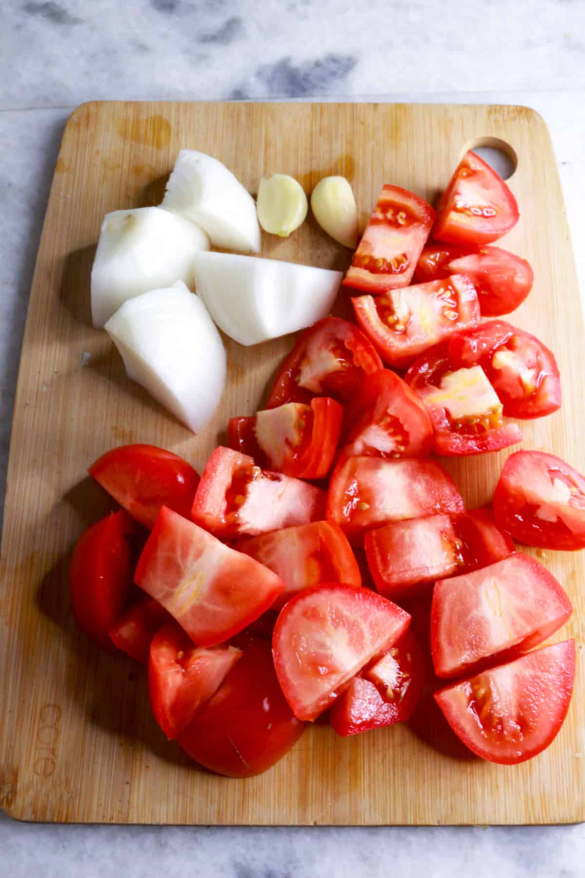 A wooden cutting board with chopped onion, chopped tomatoes and garlic cloves