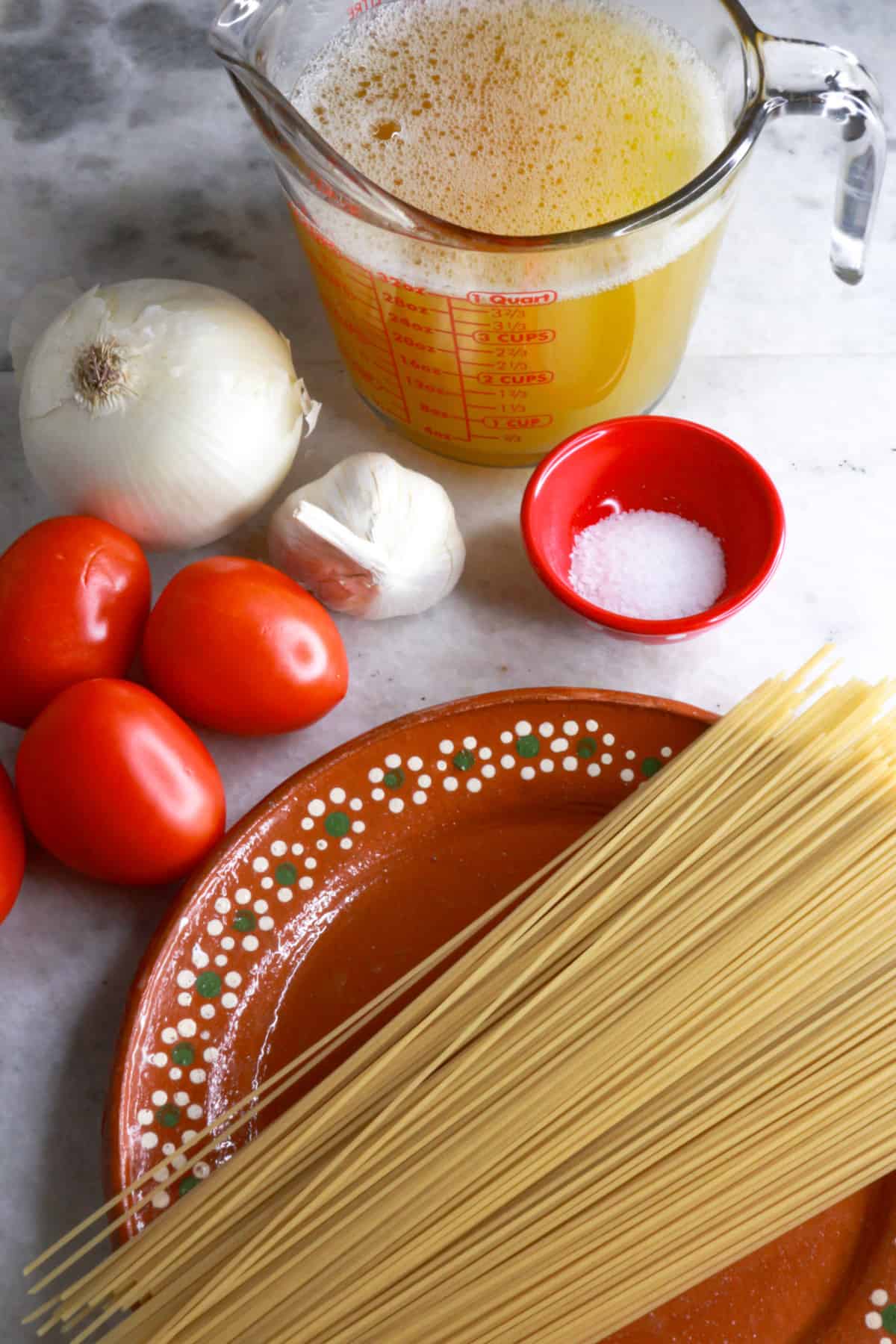 A measuring cup with chicken broth, a whole onion, 4 tomatoes, a bulb of garlic, a small bowl with salt and a plate with noodles