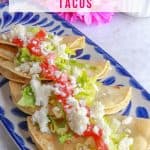 A row of crispy refried bean tacos on a platter topped with lettuce, tomatoes, and crumbled queso fresco