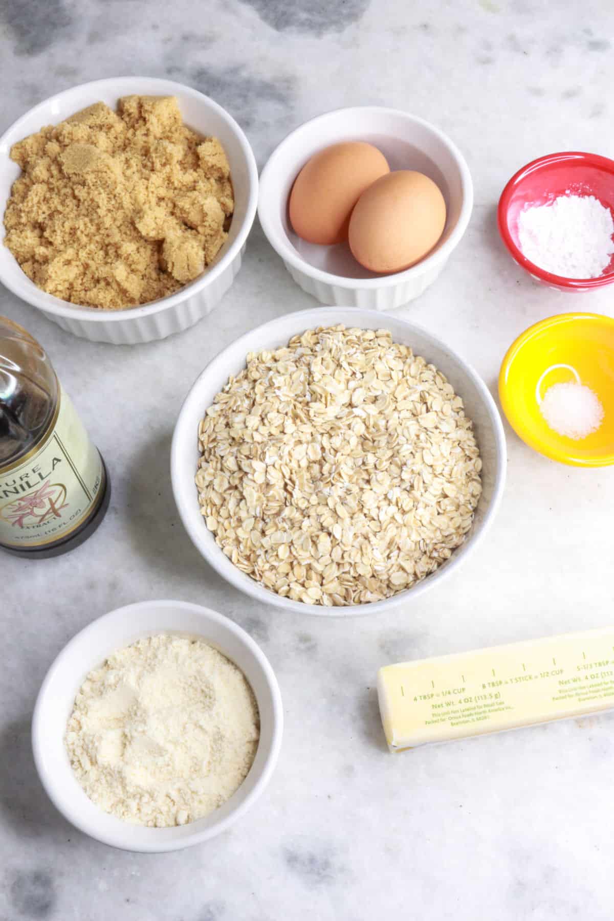 Bowl of brown sugar, 2 eggs, a pinch bowl with baking powder, another with salt, a bowl with oats, a stick of butter, a bowl with coconut flour and a bottle of vanilla extract