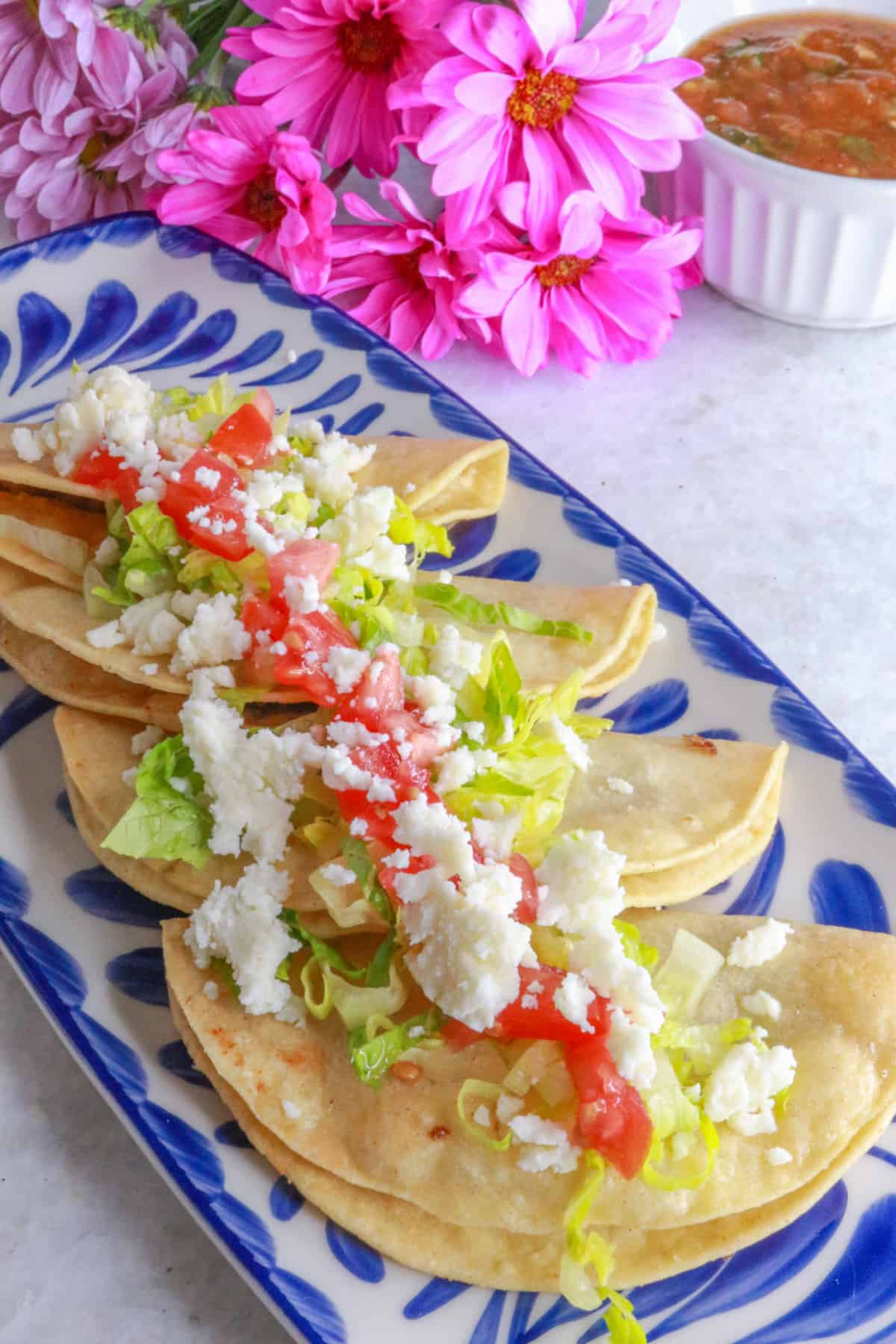 Side view of Tacos dorados de frijoles in a row on a platter, topped with lettuce, tomatoes and queso fresco