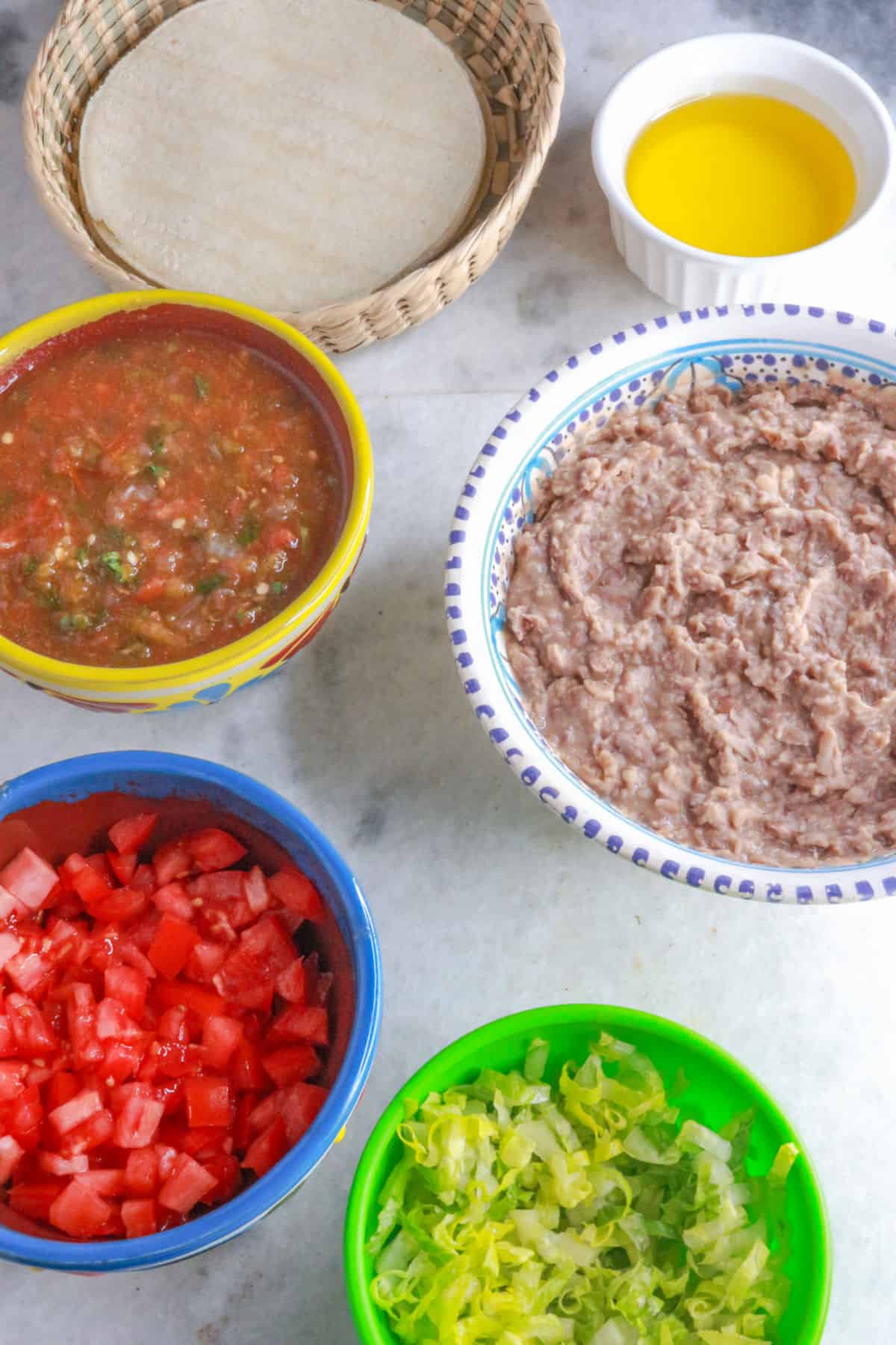 A basket with corn tortillas, a bowl with oil, a bowl with jalapeño salsa, a bowl with refried beans, another with diced tomatoes and another with shredded lettuce