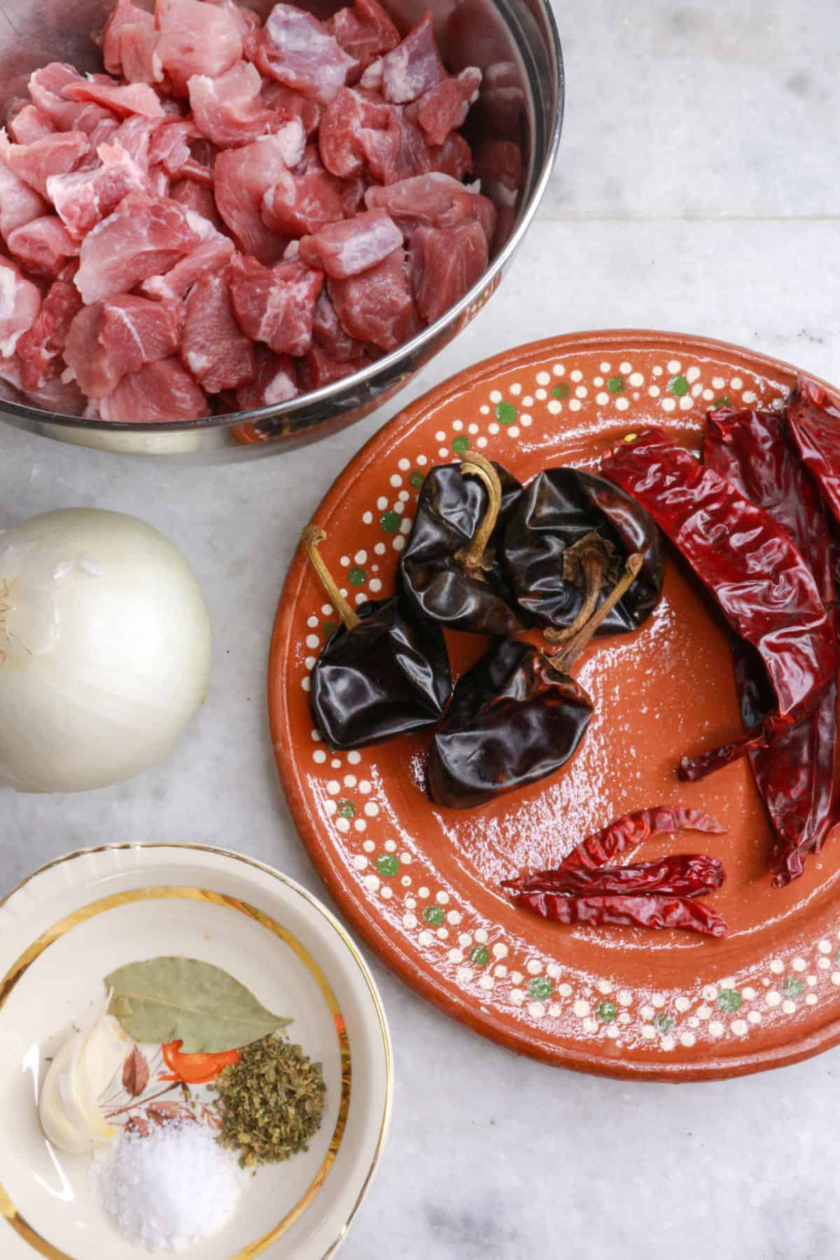 A mixing bowl with cubed raw pork, an onion, a plate with dried chiles, a small bowl with salt, oregano and a bay leaf