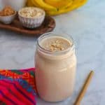 Glass filled with licuado de platano con avena (banana oatmeal smoothie) with a straw beside it and the ingredients behind it