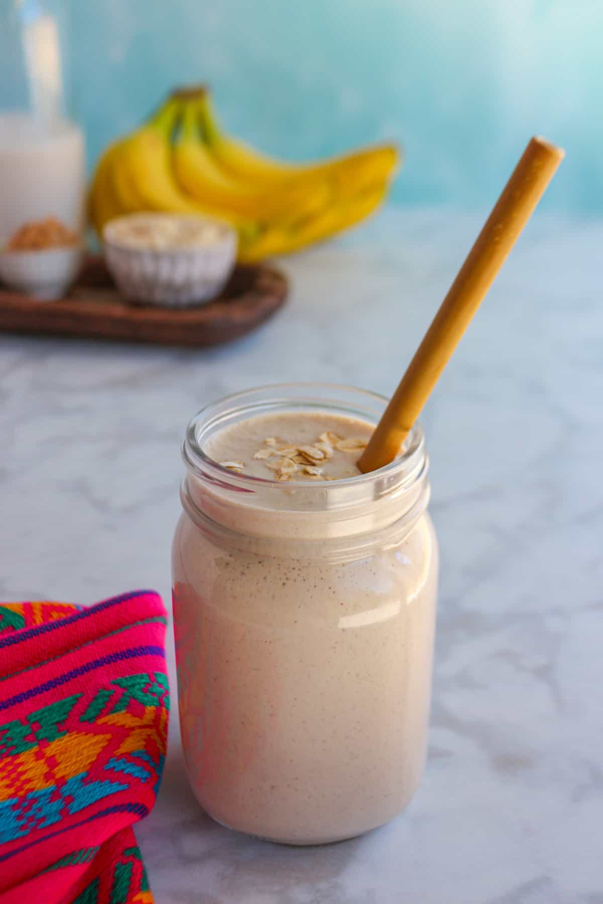 Glass filled with licuado de platano con avena (banana oatmeal smoothie) with a straw and the ingredients behind it