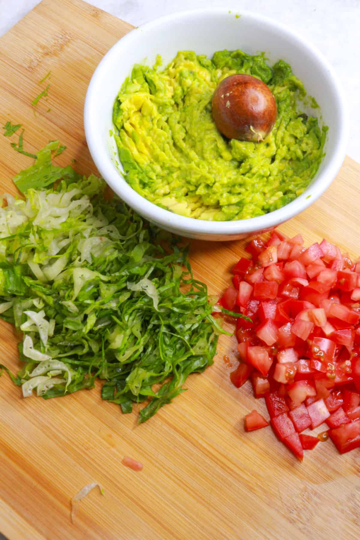 A bowl with mashed avocado on a cutting board with shredded lettuce and diced tomato