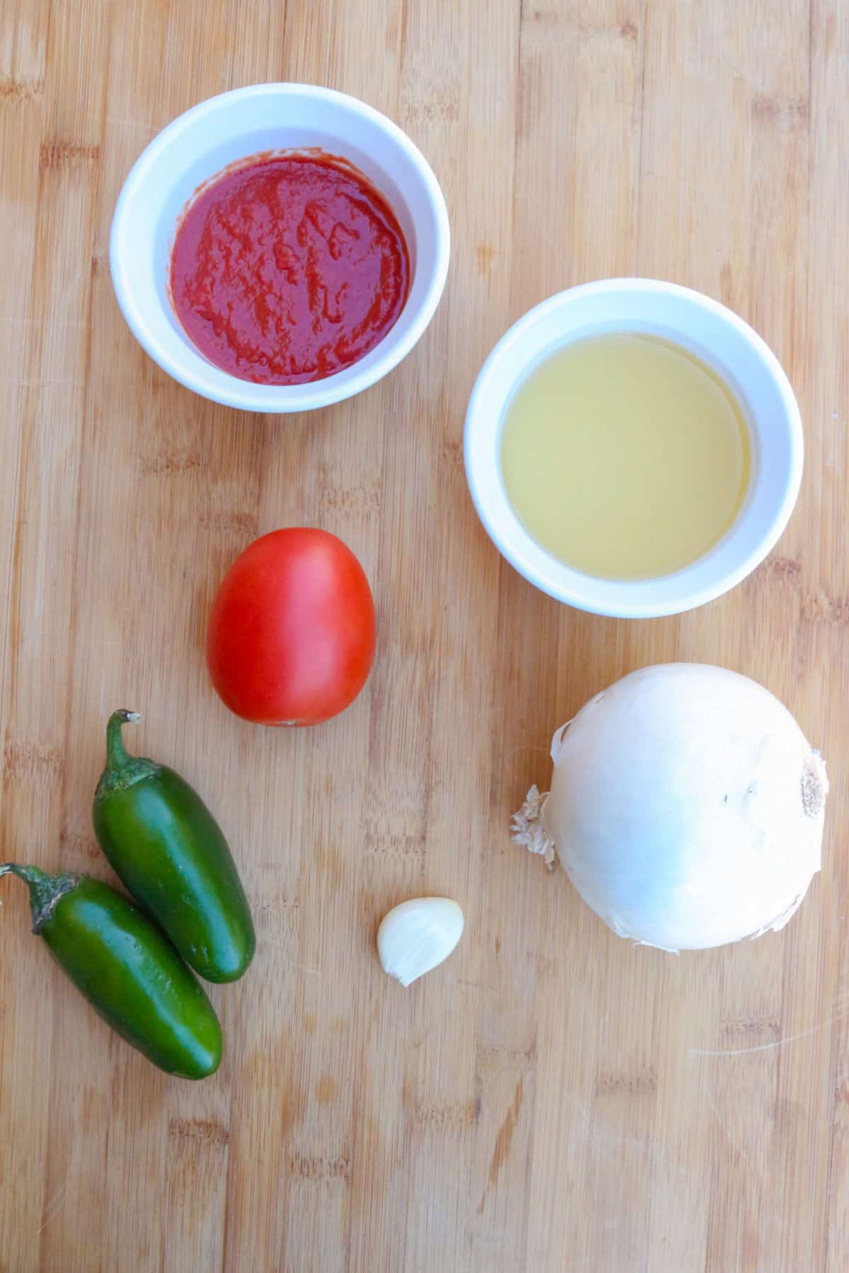 A small white bowl with tomato sauce, another with chicken broth, a Roma tomato, 2 jalapeños, one garlic clove and a white onion on a cutting board