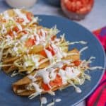 Plate of crispy tacos de papa topped with cabbage, tomatoes and sour cream