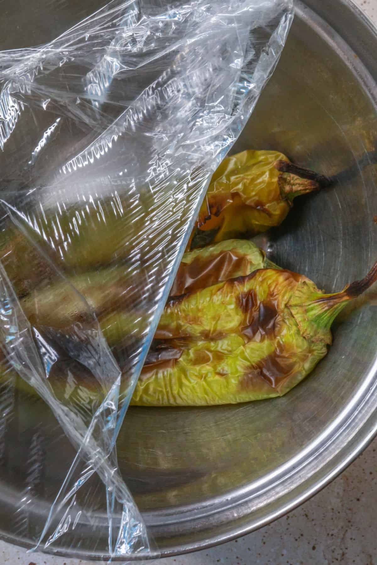 Roasted green chiles in a bowl with plastic wrap to sweat