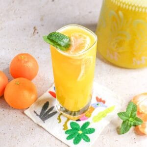 Glass of mandarin Agua Fresca with mint on a coaster with a pitcher of the Agua Fresca behind it